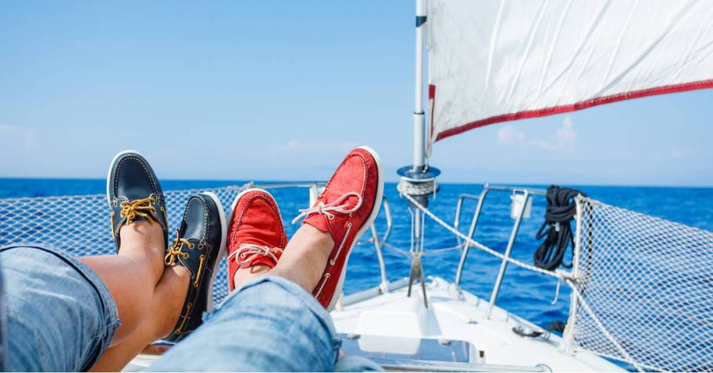  On the edge of the deck on a boat, two people sit with their legs crossed, showing off their red and blue boat shoes.