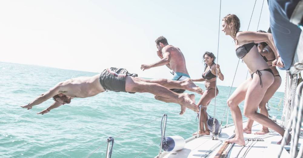 A group of people wearing swimsuits run and jump off the side of a boat toward the blueish ocean in front of them.