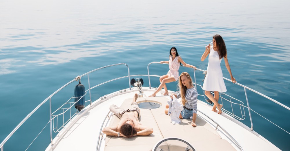 Four people are relaxing at the end of a boat, enjoying a still day on the water. The man is lying down, while the women are sitting or leaning.