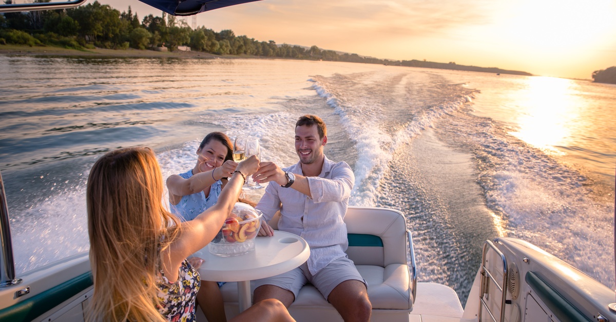 Three people sit on the back of a boat cruising down the water. They are making a toast with the sunset behind them.