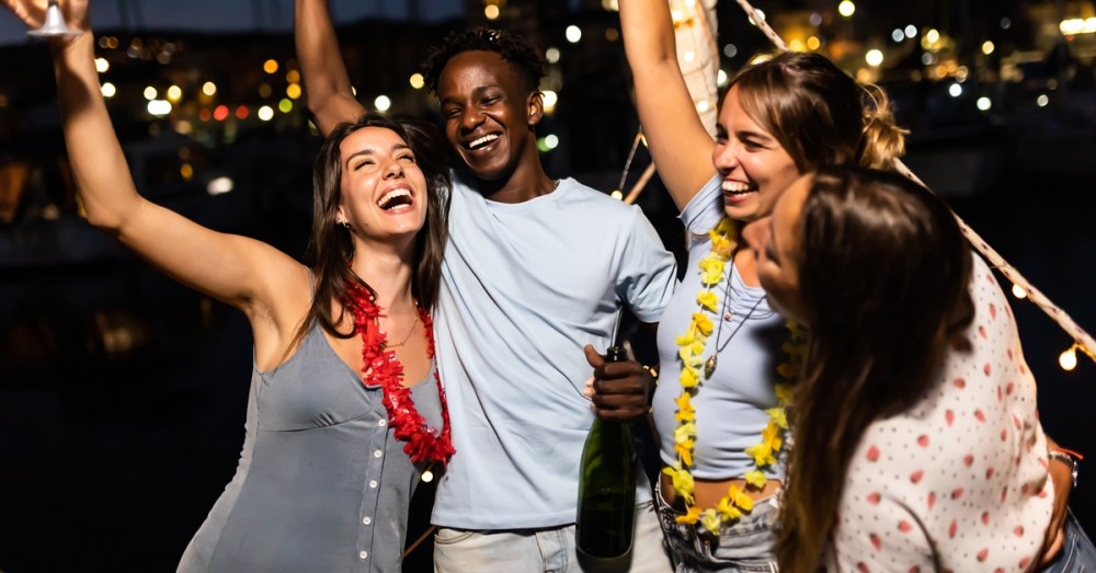 Four people on a boat smile and hold drinks up in the air. There are bright lights illuminating the night behind them.