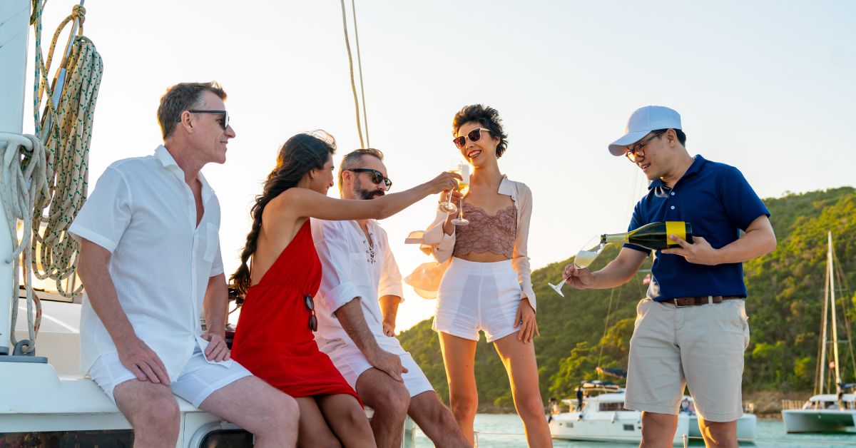 pinkpartyboat-368049-friends-boat-champagne-BlogBanner1 A few friends stand and sit together on a boat while a worker on the boat pours champagne for them.