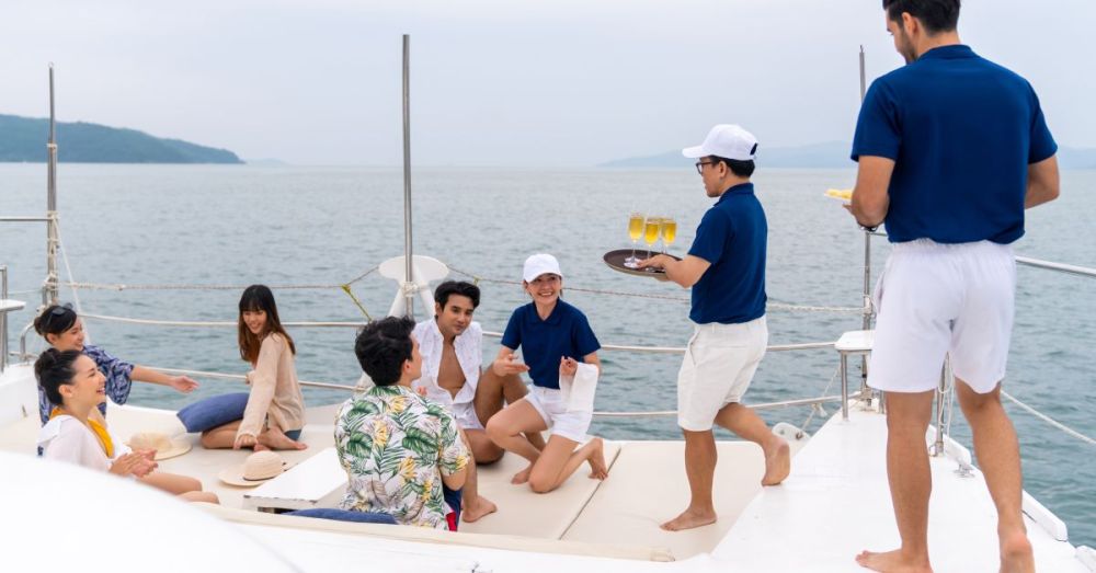 Some friends sit on a boat on the water while the boat workers walk toward them holding platters of champagne.
