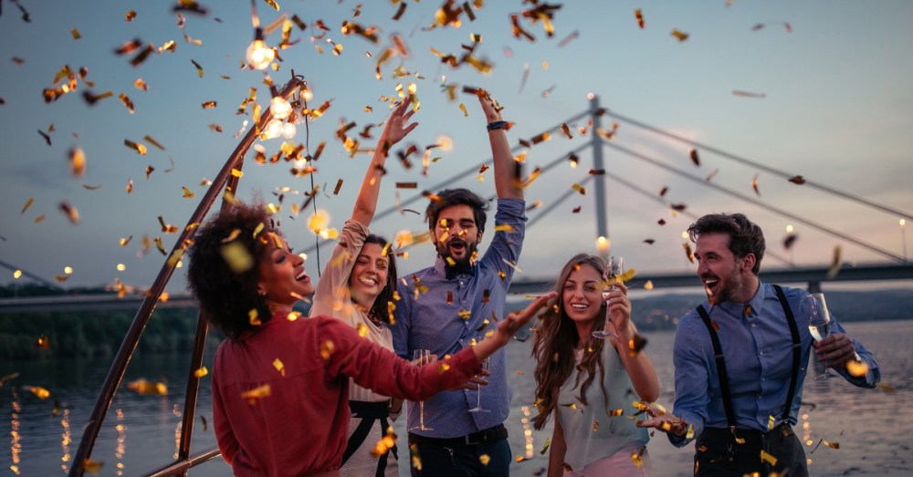 A group of friends dancing under golden confetti during an outdoor celebration with a bridge in the background.