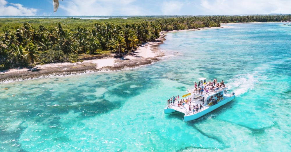 A white catamaran carrying people in swimwear while cruising near a palm-covered island with turquoise tropical water.