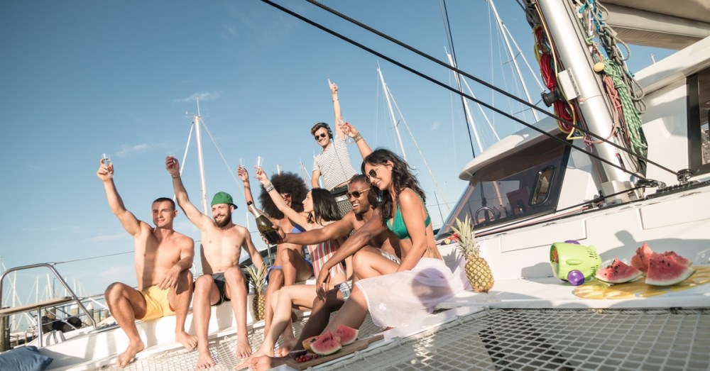 A group of people sitting on a boat, with many of them holding up glasses. There is a bowl of fruit in the foreground.