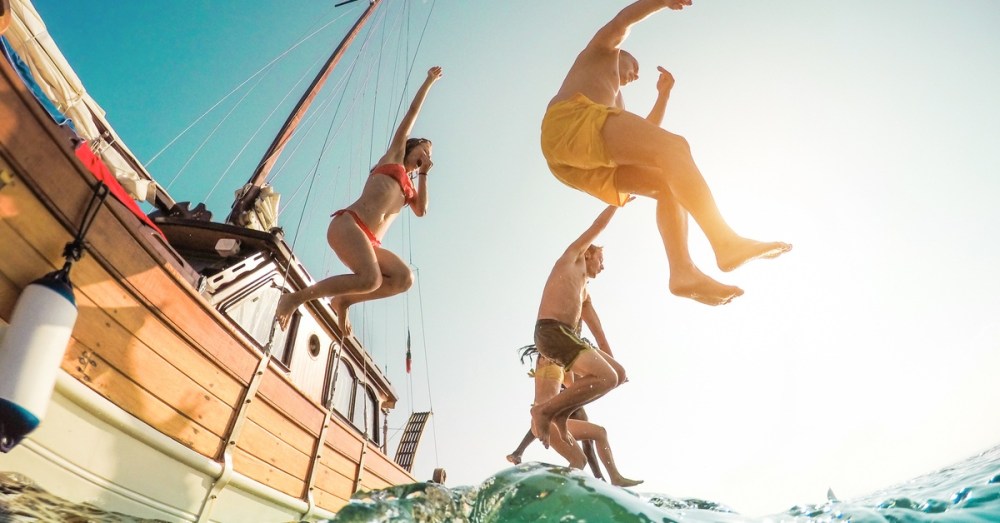 Several people jumping off a boat into the water, with some plugging their noses. There is a blue sky in the background.