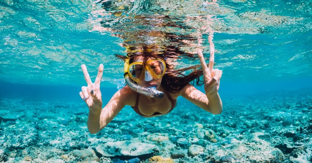 A close-up view shows a woman snorkeling in shallow water near a coral reef. She's holding up the peace sign.