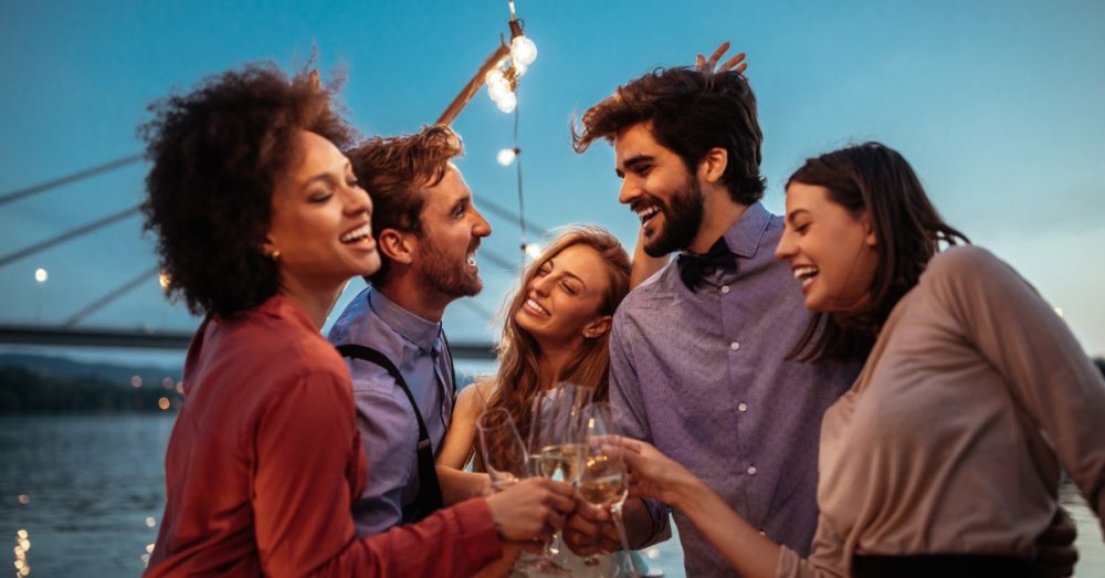 A group of three women and two men sit around on a comfy seating area on a large boat at sunset, having fun.