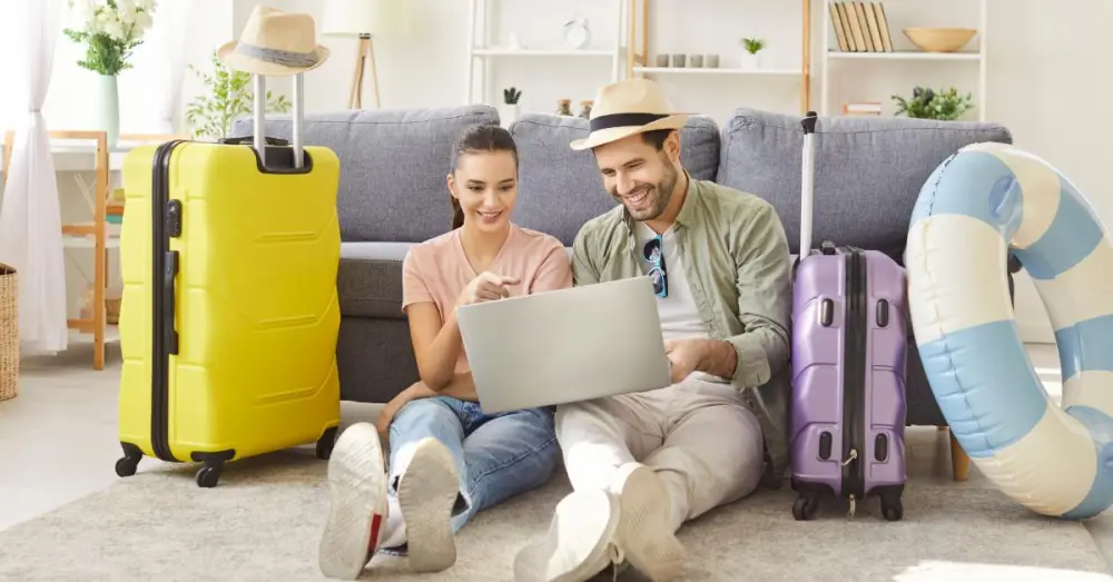 A smiling man and woman sit on the floor of a living room looking at a laptop. Luggage sits next to them.