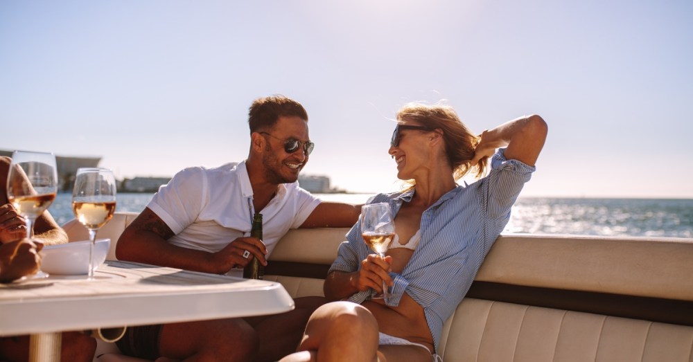 A man and a woman sit on a leather seat on a boat in front of a table, having a drink and smiling at each other.
