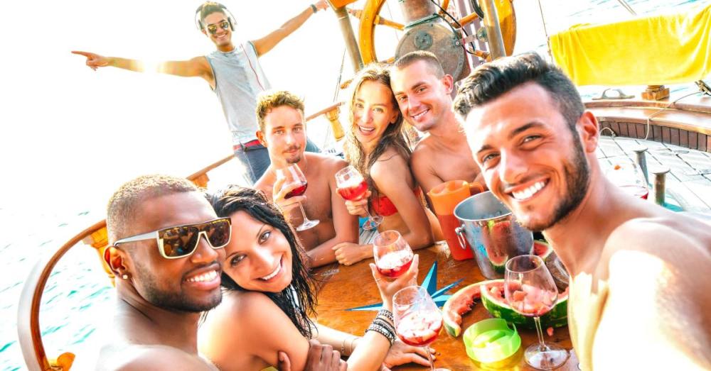 Six people sit around a table that's on a boat, holding drinks and posing for a photo. A person stands in the background.