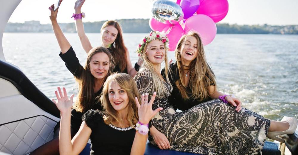 Five women pose for a photo at the back end of a boat. A balloon bouquet of pink and purple balloons is next to them.