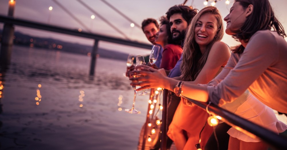 Five people lean on the railing of a boat while holding various alcoholic beverages. The railing has string lights.