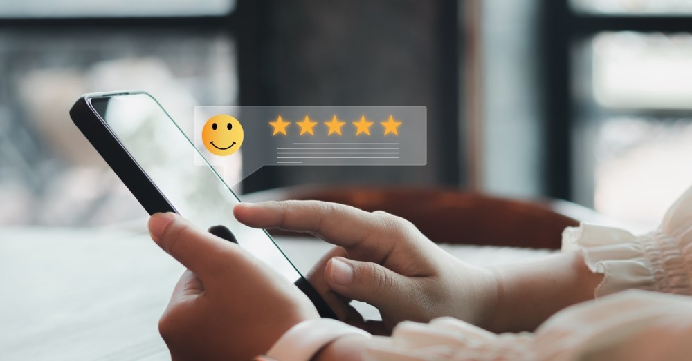 A hand holds a smartphone displaying a five-star rating and smiley review icon against a softly blurred interior.