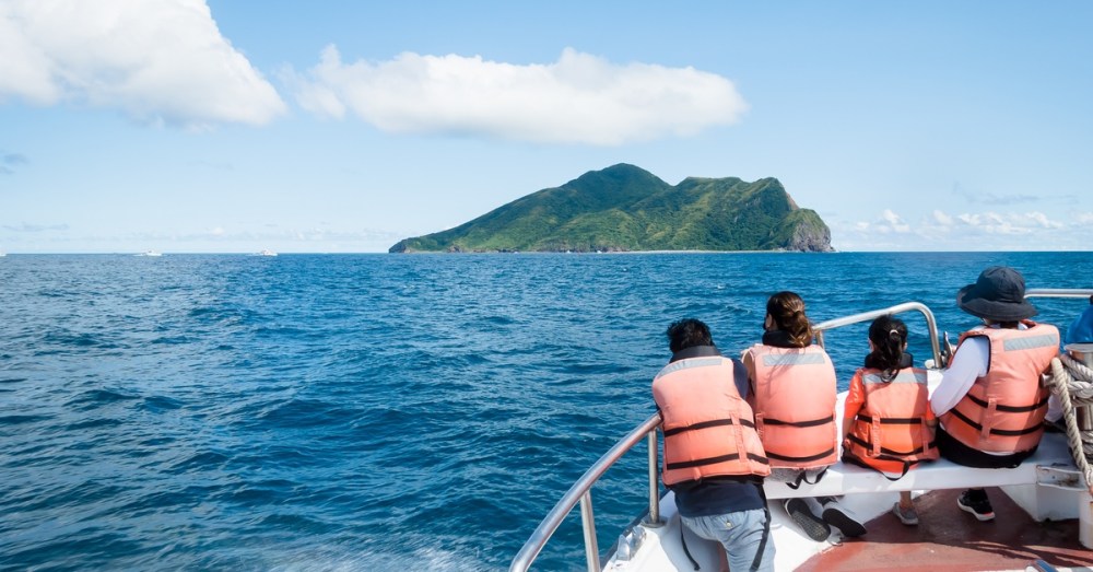 Four passengers in orange life vests sit at the bow of a speedboat, facing a green island across bright blue water.