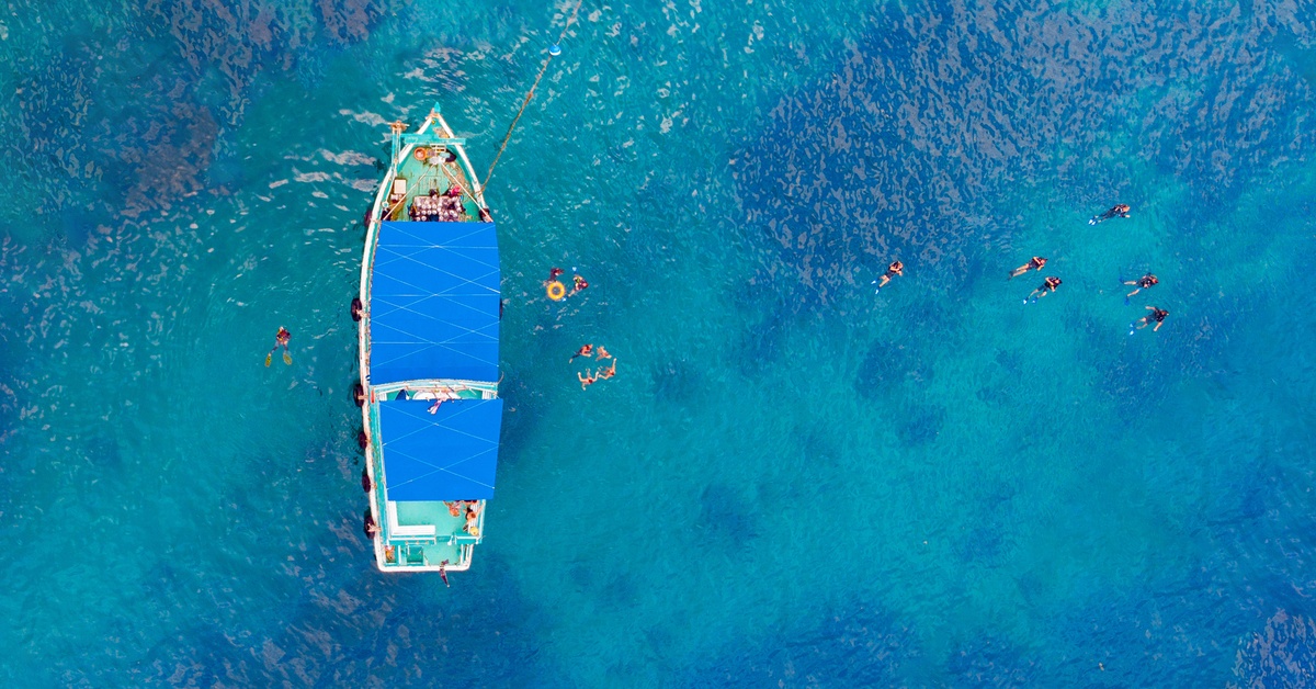 An aerial view of a tour boat anchored in clear turquoise water, with scattered swimmers floating nearby.