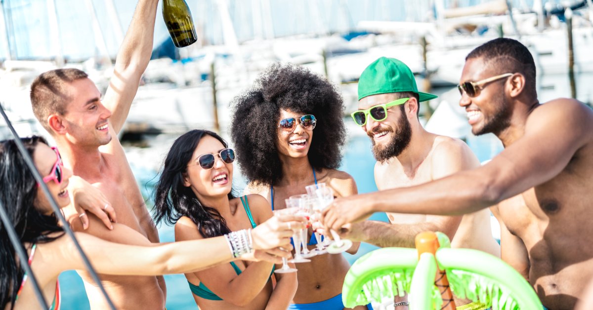 Three men and three women gather around a small inflatable palm tree to toast with small, clear glasses on a boat.