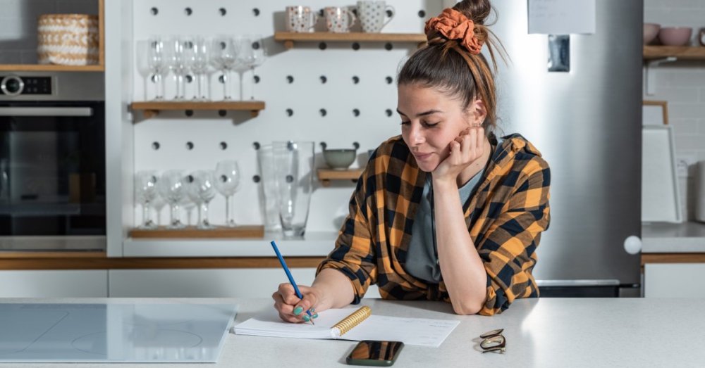 A young woman leans on a white kitchen counter while resting her chin on one hand and writing in a notepad with the other.