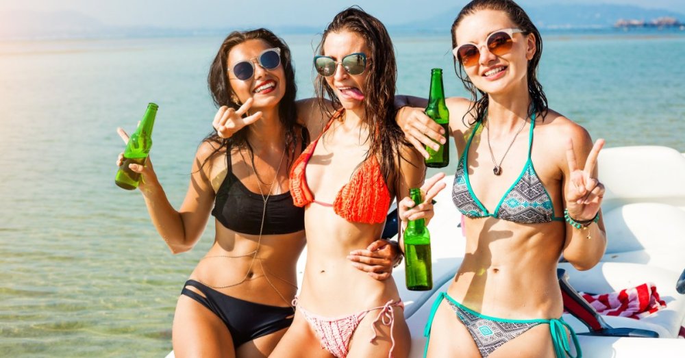 Three women wearing bathing suits pose for a photo while holding beers. They are smiling and making peace signs.
