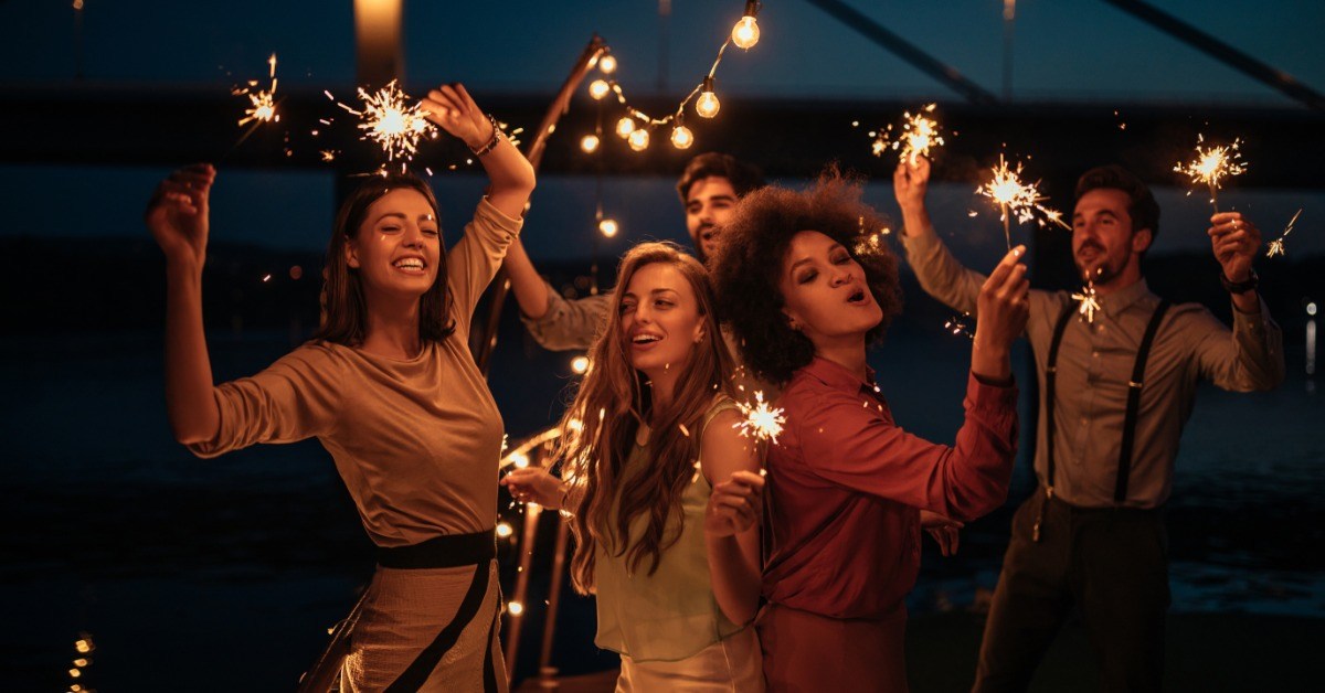 Three women and two men are dancing on a party boat at night. Each person is holding a lit sparkler.
