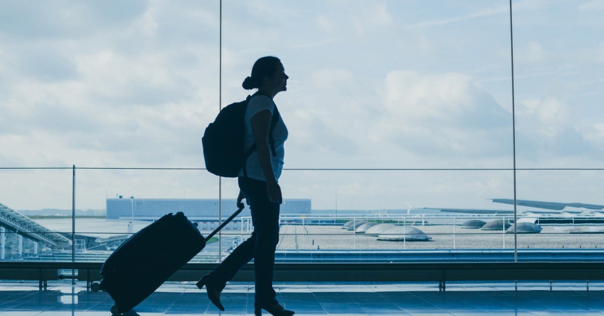 A woman is walking in an airport next to a large window carrying a backpack and pulling a suitcase.