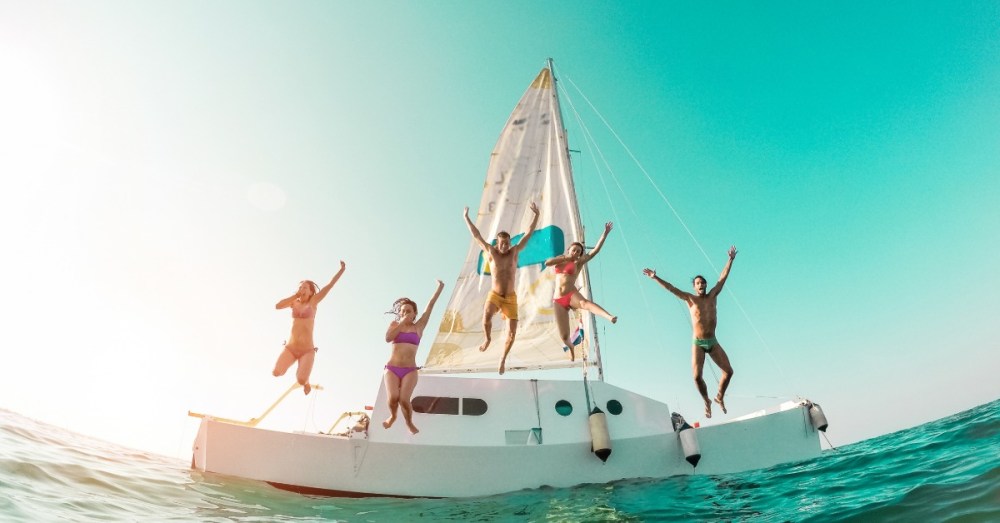Three women and two men are jumping off the side of the boat into the body of water. The blue sky is behind them.