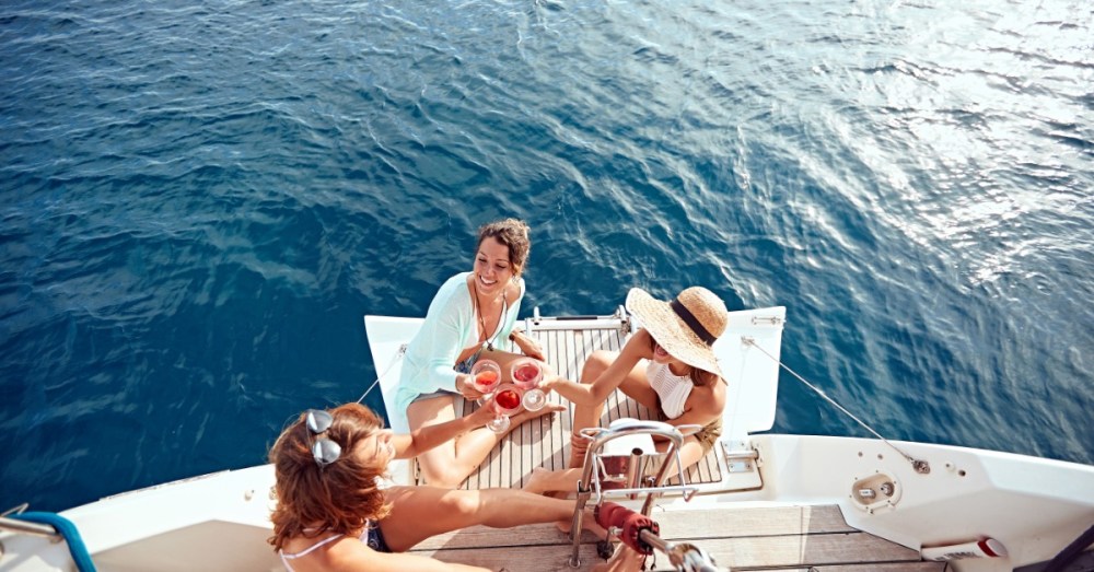 An aerial view shows three women sitting on the back of the boat, placing their glasses into the center for a cheers.